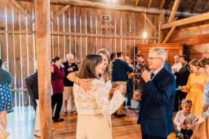 People socialize in a rustic, wooden venue; two women with drinks smile and talk with an older man, while others mingle in the background.