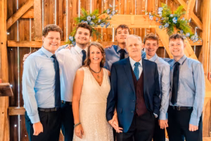 A group of six men in dress shirts and ties and one woman in a cream dress pose together in front of a wooden backdrop decorated with blue flowers.