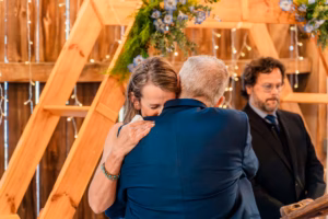 A woman and an older man embrace closely at a formal event, while another man stands nearby in front of wooden beams and decorative flowers.