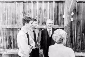 Four people, three men in dress shirts and ties and one woman with short hair, stand and talk in front of a wooden wall with string lights.