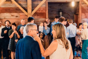 A woman in a white dress gently touches a man's face while a group of people gathers and chats in the background inside a rustic wooden venue.