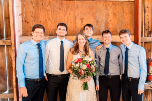 A woman in a white dress holding a bouquet stands with five young men in blue shirts and dark ties in front of a wooden wall.