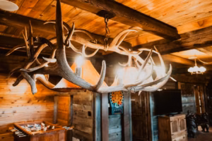 A rustic chandelier made of antlers hangs from a wooden ceiling in a cabin, with warm lighting and wooden furnishings visible in the background.