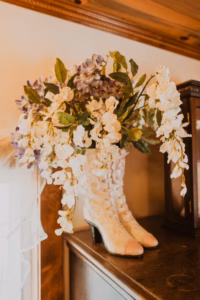 A pair of vintage white lace-up boots used as a vase, holding a bouquet of white and purple flowers, displayed on top of a wooden surface next to a wooden cabinet.