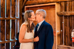 A woman in a white dress kisses an older man in a suit on the lips inside a wooden barn.