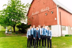 Six men in dress shirts and ties stand smiling in front of a red barn labeled "Justin Trails" on a grassy lawn with trees in the background.