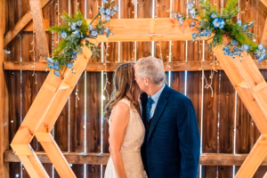A couple in formal attire shares a kiss under a wooden hexagonal arch decorated with greenery and blue flowers, in front of a rustic wooden backdrop.