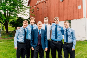 Six men, dressed in formal and semi-formal attire with blue shirts and ties, stand together smiling in front of a red barn on a grassy lawn.