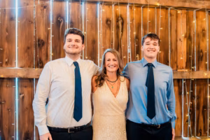 Three people, two men in dress shirts and ties and a woman in a white dress, stand smiling in front of a wooden wall with string lights.