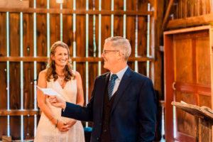 A man in a suit speaks while holding a piece of paper; a smiling woman in a white dress stands beside him in a rustic wooden setting.