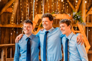 Three young men wearing blue shirts and dark ties stand together with arms around each other, smiling, in front of a wooden backdrop with string lights.