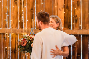 A woman smiles while hugging a man who holds a bouquet of flowers, standing in front of a wooden wall decorated with string lights.