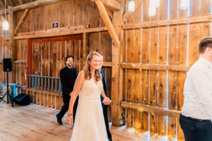 A woman in a white dress walks hand-in-hand with a man in a suit inside a wooden venue decorated with string lights. Two other people are visible in the background.