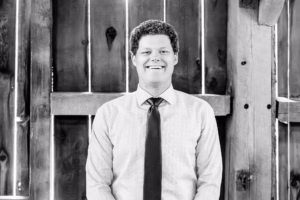 A man in a dress shirt and tie stands smiling in front of a wooden wall. The photo is black and white.