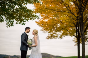 A bride and groom stand facing each other outdoors, holding a bouquet, with autumn trees and cloudy skies in the background.