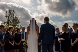 A bride and groom stand holding hands outdoors, facing a group of people who are arranged in a semicircle under a partly cloudy sky.