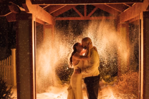 A couple in formal attire kisses under a wooden pergola at night, surrounded by falling snow illuminated by warm lights.