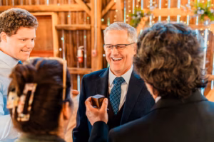 Four people stand in a warmly lit indoor setting; one man in a suit is smiling while another person holds a small wooden box toward him.
