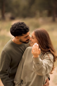 A couple stands close together outdoors; the woman smiles and shows her hand with a ring, while the man leans in affectionately from behind.