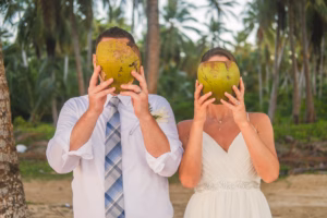 A couple in wedding attire stands outdoors holding large coconuts in front of their faces, with palm trees in the background.