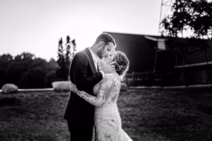 A bride and groom in formal wedding attire share a kiss outdoors, with trees and a building in the background, captured in black and white.
