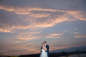 A bride and groom stand embracing outdoors at sunset with a dramatic, cloud-filled sky in the background.