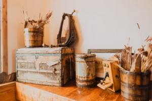 A collection of vintage wooden barrels, an old trunk, a mirror, and an antique sewing machine arranged on a wooden floor against a plain wall.