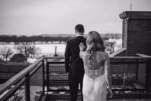 A bride in a strapless dress approaches a groom from behind on a balcony, reaching out to touch his shoulder; landscape and trees are visible in the background.