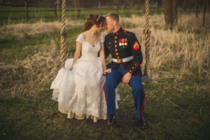 A bride in a white wedding dress and a groom in a military uniform sit closely together on a rustic rope swing in an outdoor grassy area.