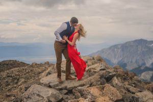 A couple stands on rocky terrain at a mountain summit, with the woman in a red dress and the man in formal attire, sharing a kiss under a cloudy sky.