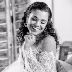 A woman in a lace wedding dress smiles with her eyes closed, resting her chin on her hand in a relaxed pose.