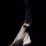 A person in a lace dress is partially illuminated against a dark background, holding the bottom of the dress with one hand.