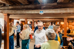 A group of people stand and converse in a rustic room with wooden beams and columns. Some hold drinks, while others talk in small groups throughout the space.