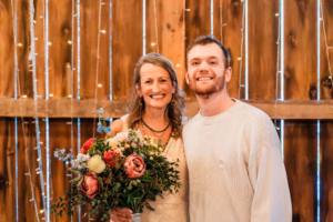Two people stand smiling in front of a wooden backdrop with string lights; the woman holds a bouquet of flowers.