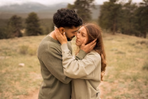 A couple stands outdoors, facing each other and embracing, with trees and mountains in the blurred background.