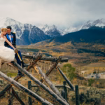 A bride and groom sit on a wooden fence overlooking a scenic mountain landscape with snow-capped peaks and autumn colors.