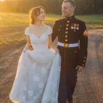 A bride in a white lace gown walks hand-in-hand with a groom in a formal military uniform on a dirt road at sunset.