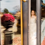 A bride in a white gown is seen through a glass door, with her reflection also visible, and a large planter with red flowers outside.