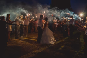 A bride and groom stand in the center of a path at night, surrounded by guests holding lit sparklers as smoke drifts through the air.