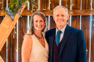 A woman in a white dress and a man in a dark suit with a blue tie stand together smiling in front of a wooden backdrop decorated with string lights and greenery.
