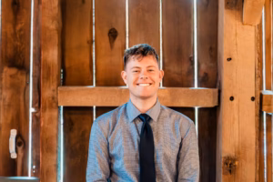 A person in a gray shirt and black tie smiles while sitting in front of a wooden wall with vertical planks and light shining through gaps.