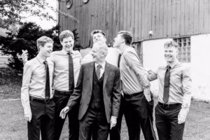 A group of six men, dressed in shirts and ties, stand together outdoors, smiling and laughing in front of a wooden building.