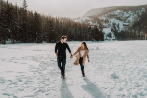 A man and woman walk hand in hand through a snowy landscape with pine trees and mountains in the background.