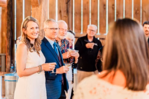 A woman in a white dress and a man in a blue suit stand together holding drinks, smiling, while people in the background also hold drinks and face a woman in the foreground.