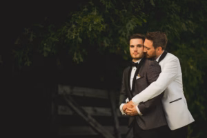 Two men dressed in tuxedos embrace outdoors; one wears a white jacket and hugs the other from behind against a dark, leafy backdrop.