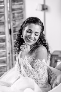 A woman in a lace wedding dress sits and smiles with her eyes closed, resting her chin on her hand.