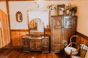 A vintage wooden vanity with mirror and an ornate wardrobe stand against a wood-paneled wall, next to a round-backed chair and decorative flowers.