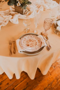 A round table set with a floral plate, utensils, two glasses, and a napkin on a cream tablecloth, with a flower centerpiece and decorative fabric runner.