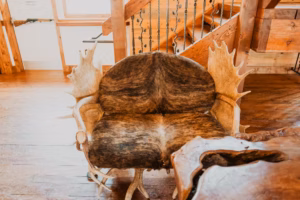 A chair upholstered with animal fur and featuring large antlers for armrests sits on a wooden floor near a staircase with wrought iron railing.