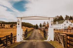 A paved driveway leads through a white gate marked "Deer Creek Valley Ranch," flanked by wooden fences, with a large white barn and trees in the background.